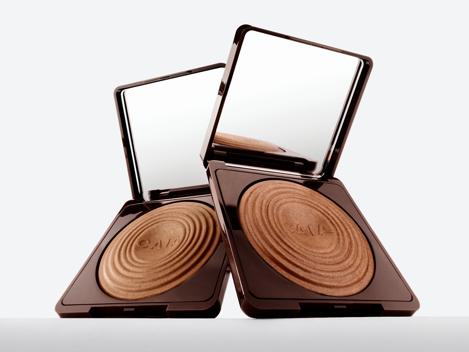 Zwei quadratische Make-up-Kompaktpuder mit Spiegeln in braunen Gehäusen, die einen schimmernden Bronzeton mit einem gewellten Design enthüllen.