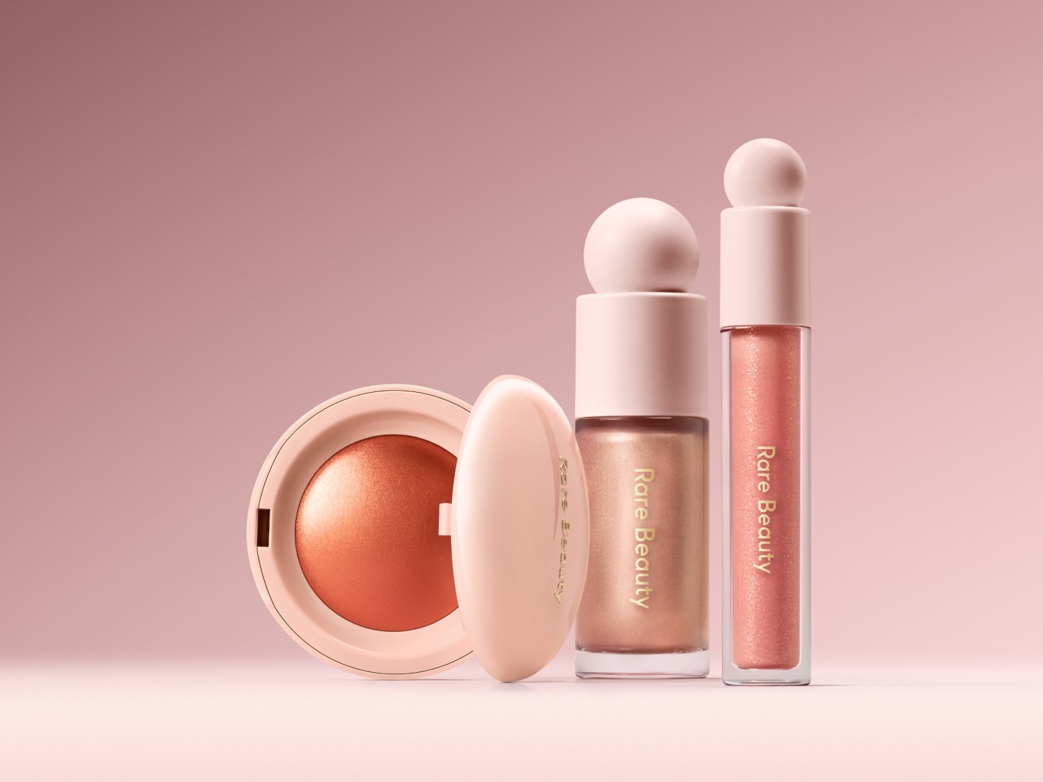Een close-up van Rare Beauty make-up producten in zachte, roze tinten, waaronder blush, vloeibare highlighter en lipgloss, allemaal gepresenteerd op een bijpassende roze achtergrond.
