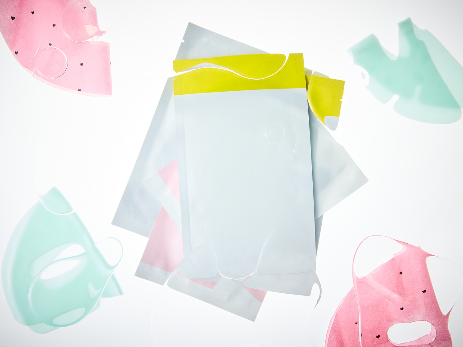 Verschiedene Tuchmasken in Rosa- und Grüntönen, einige mit Herzchenmuster, liegen neben weißen und gelben Verpackungen.