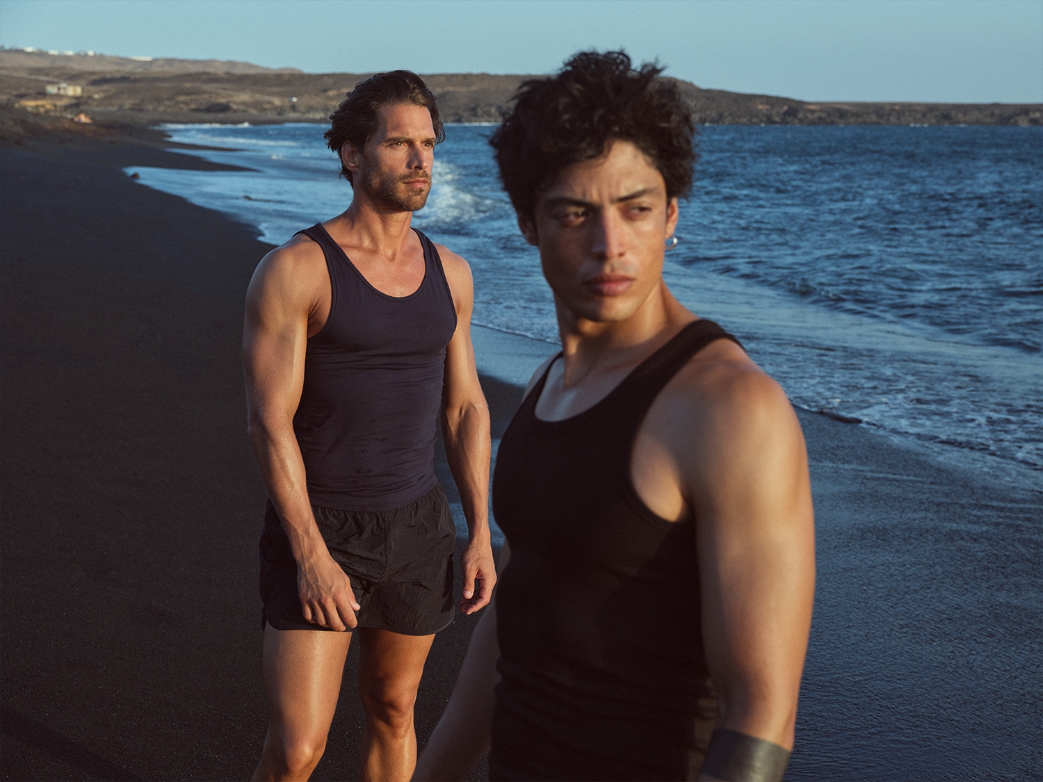 Zwei muskulöse Männer in schwarzen Tanktops und Shorts stehen an einem dunklen Sandstrand mit dem Meer im Hintergrund.