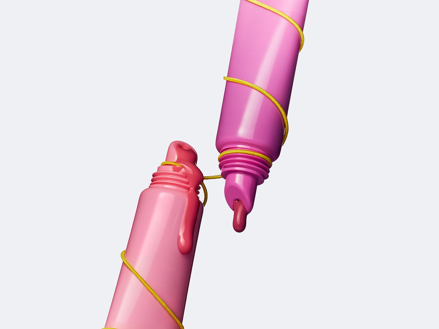 Zwei rosa Lipgloss-Tuben, verbunden mit einem goldenen Faden, wobei aus beiden Tuben etwas von dem glänzenden, farbigen Inhalt austritt.