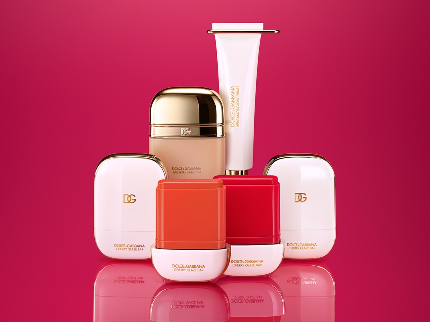 Eine Auswahl von Dolce & Gabbana Make up Produkten, darunter Cherry Glaze Bar in Rot- und Orangetönen, eine Blueberry Nutri-Tint Foundation und Bergamot Glow Primer, vor einem rosa-roten Hintergrund.