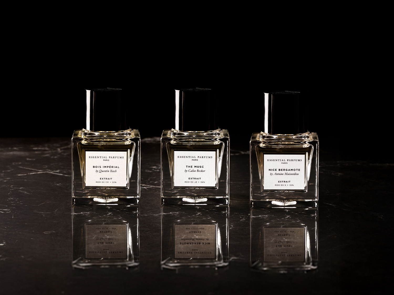 Drei quadratische Parfümflaschen von Essential Parfums stehen auf einer glänzenden Oberfläche, jede mit einem Etikett, das die Duftrichtung und den Namen des Parfümeurs angibt.