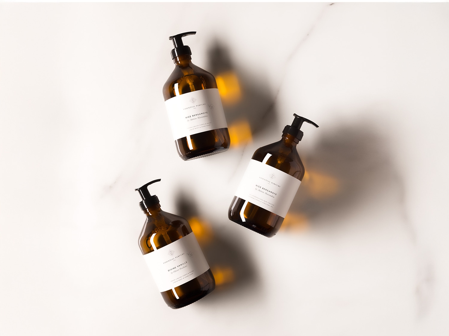 Drei braune Glasflaschen Essential Parfums mit Pumpspender, beschriftet mit "Nice Bergamote" und "Divine Vanille" auf einem weißen Marmorhintergrund.
