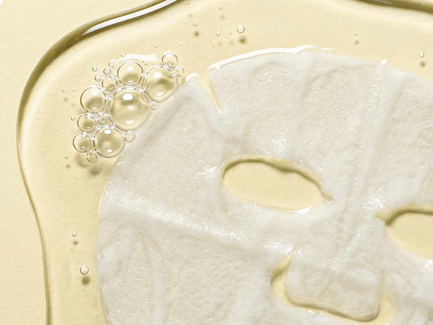 Een wit gezichtsmasker doordrenkt met serum, omgeven door heldere bubbels en een glanzende, lichtgele vloeistof.