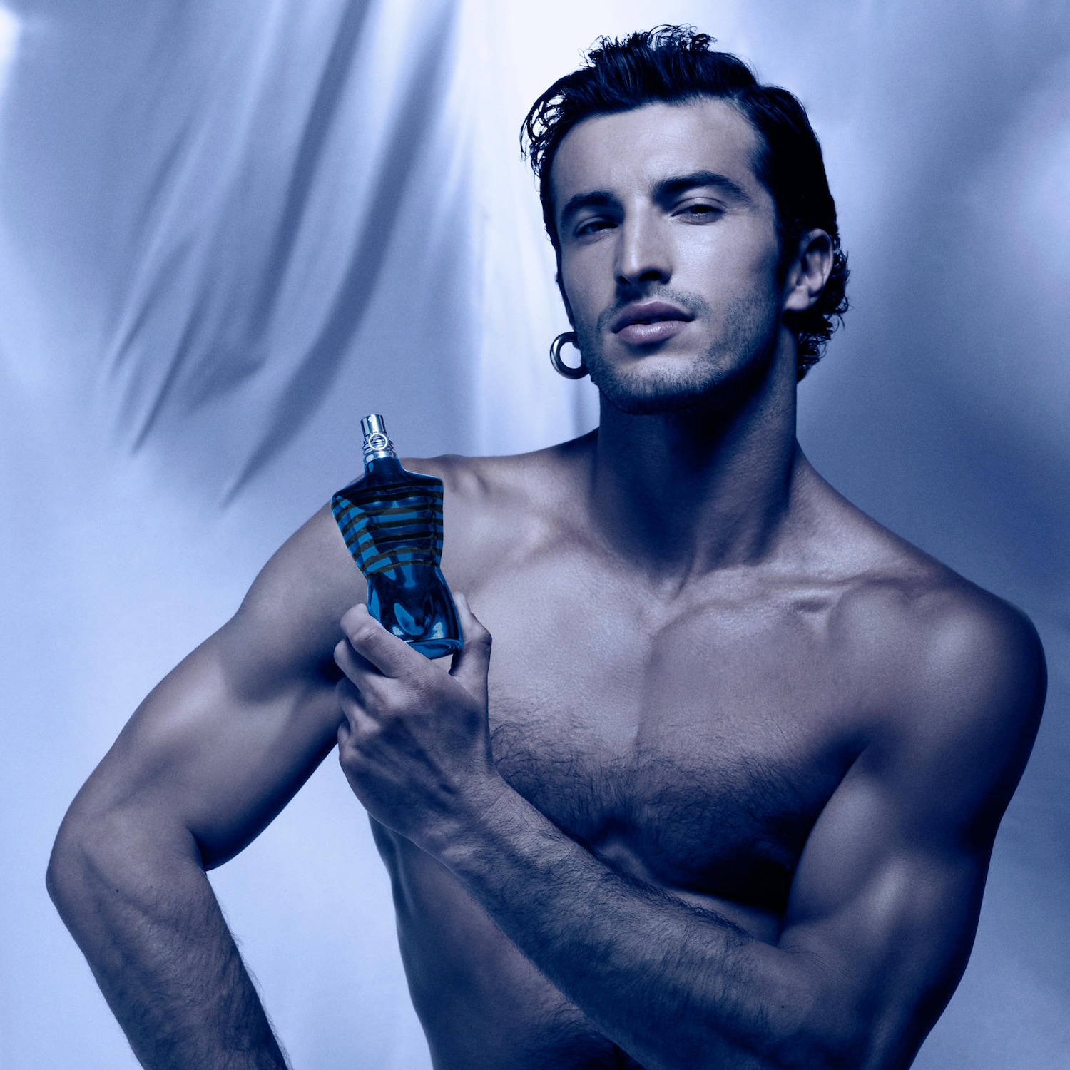 Un homme torse nu tient un flacon de parfum bleu en forme de torse masculin rayé, évoquant une allure à la fois fraîche et séduisante.
