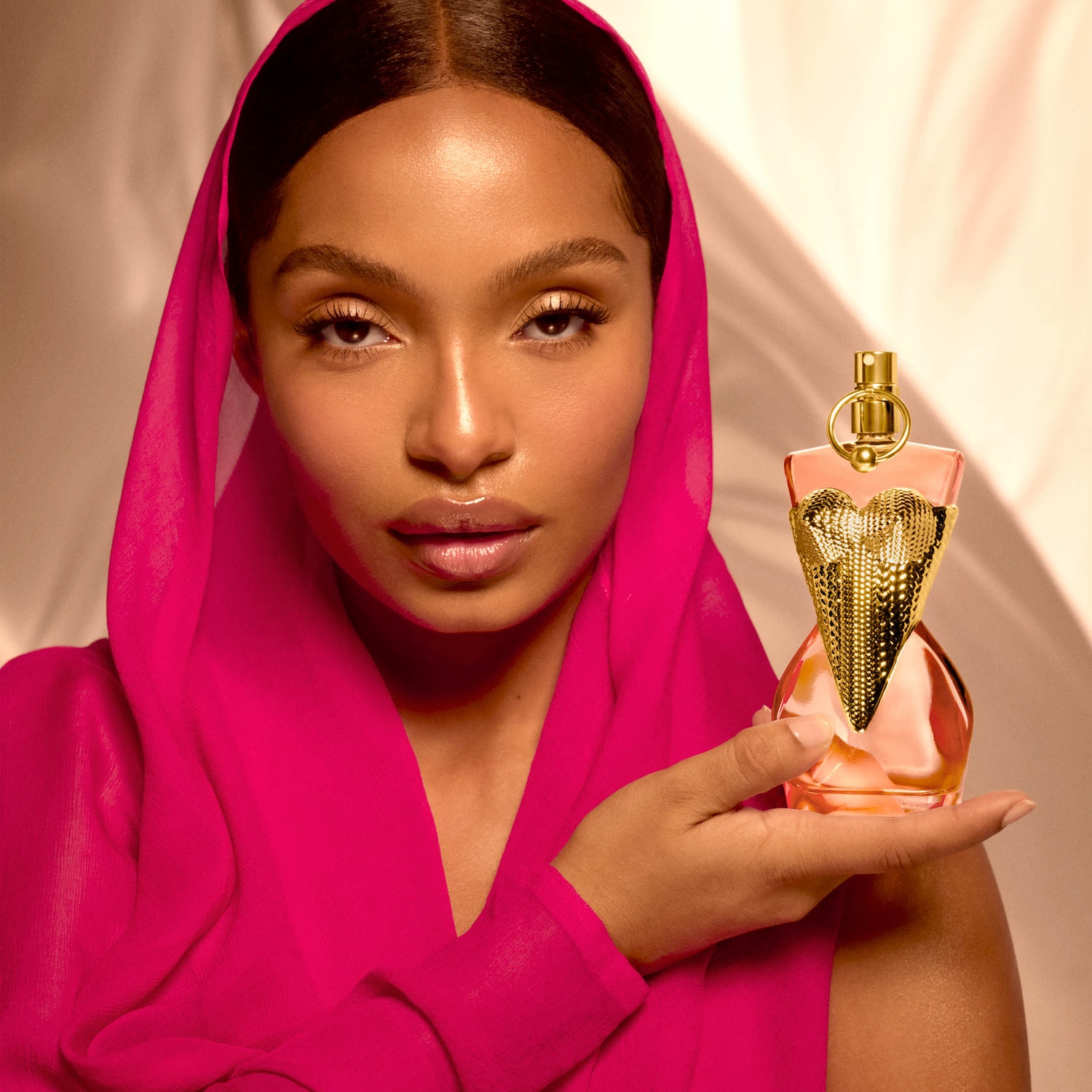 Une femme portant un foulard rose vif tient un parfum dans un flacon rose en forme de corps de femme avec des détails dorés.