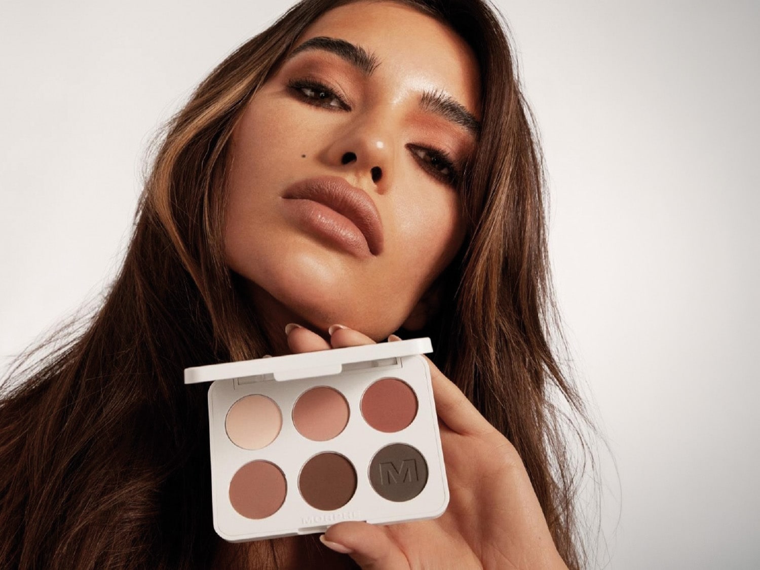 Una donna con lunghi capelli castani tiene in mano una palette di ombretti Morphe bianca con sei tonalità di marrone e rosa, mostrando un trucco occhi e labbra coordinato.