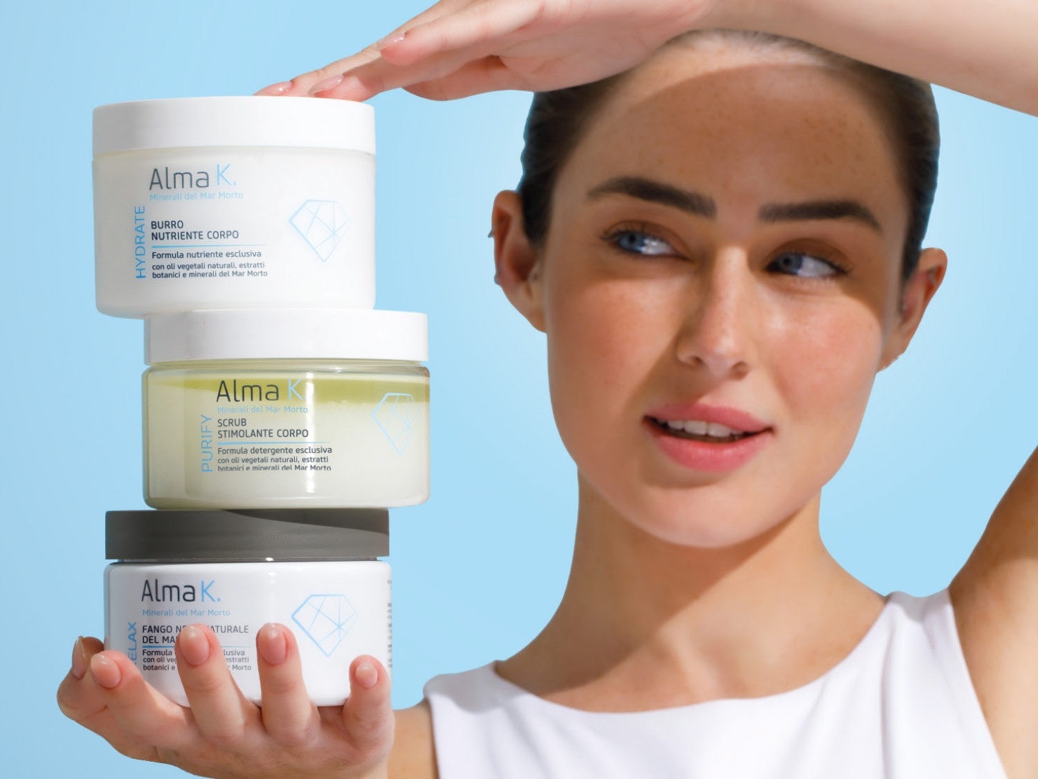 Una donna sorridente tiene in mano tre vasetti di prodotti Alma K. Minerali del Mar Morto: un burro nutriente corpo bianco, uno scrub stimolante corpo verde chiaro e un fango naturale corpo grigio, tutti con etichette che descrivono i loro benefici.
