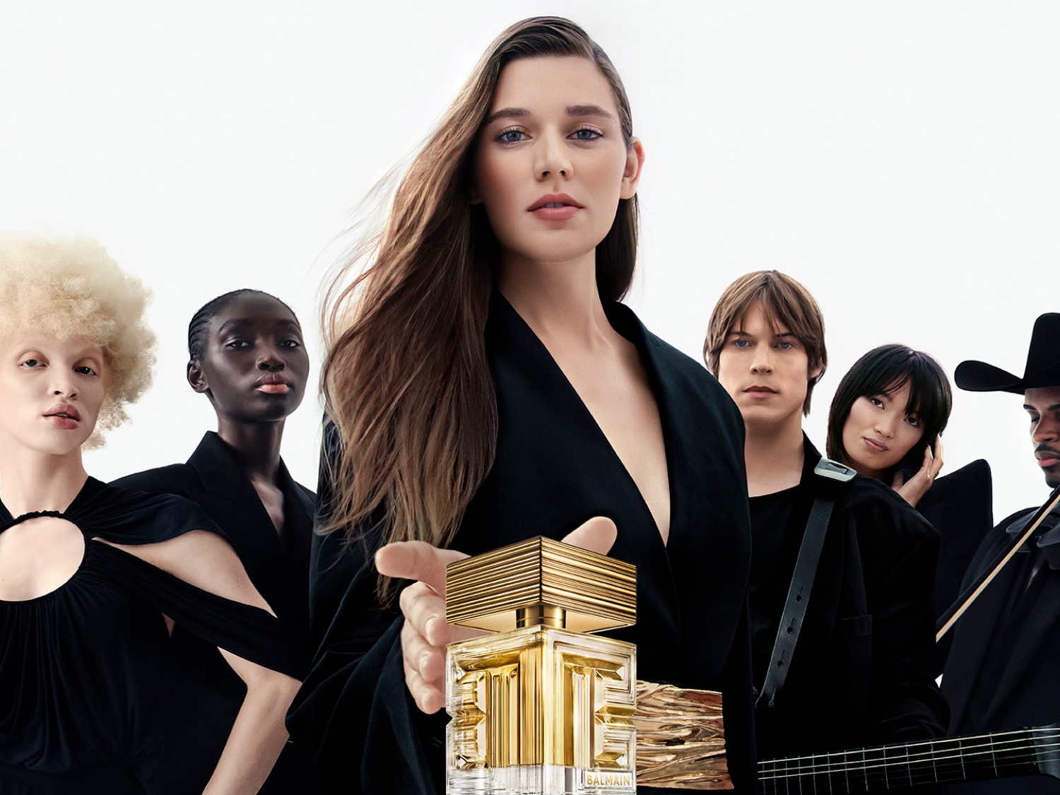 Una donna con lunghi capelli castani tiene in mano una bottiglia di profumo Balmain dorata, circondata da un gruppo diversificato di persone, tutte vestite di nero su sfondo bianco.