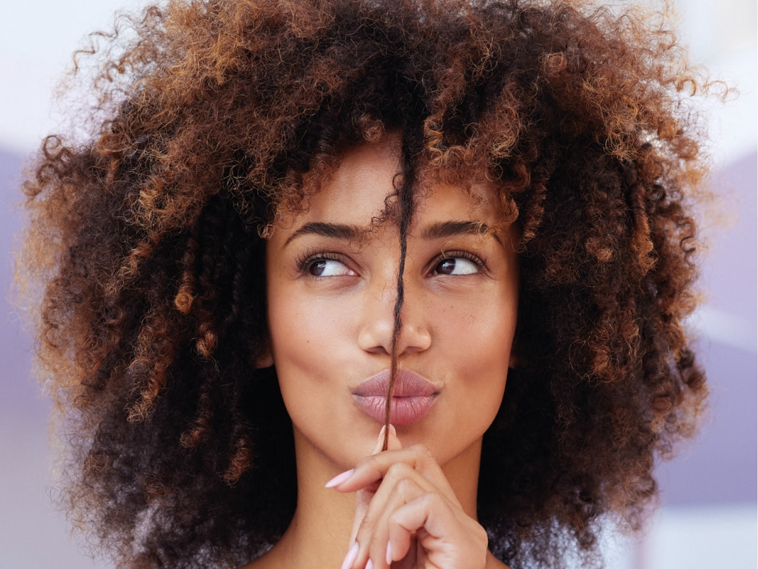 Una donna con capelli ricci castani e biondi, con una ciocca di capelli che le divide il viso, guarda di lato con un'espressione pensierosa.