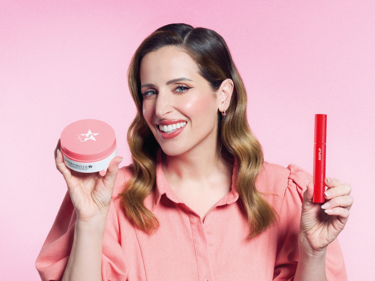 Una donna sorridente con capelli castani mossi e una camicia rosa, tiene in mano un barattolo rosa e bianco di Superstrucco e un mascara rosso 360Flip.