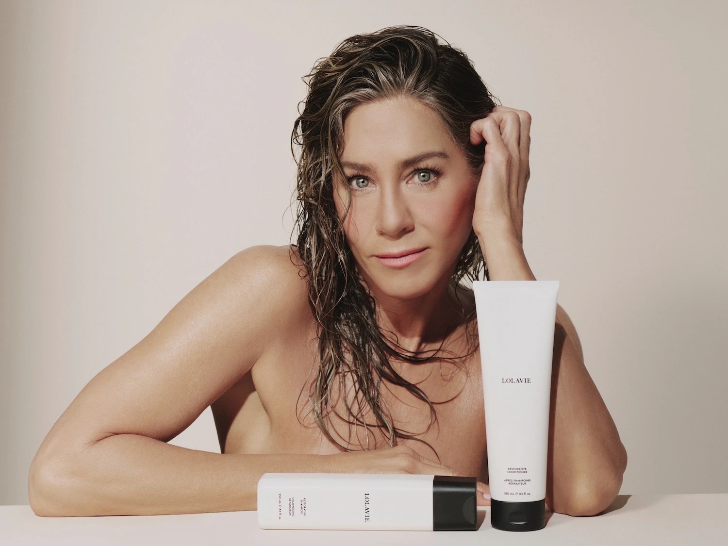 Jennifer Aniston, con i capelli bagnati e un'espressione naturale, presenta due prodotti per capelli LolaVie: un flacone bianco con tappo nero e un tubetto bianco con tappo nero, entrambi con il logo LolaVie in nero.