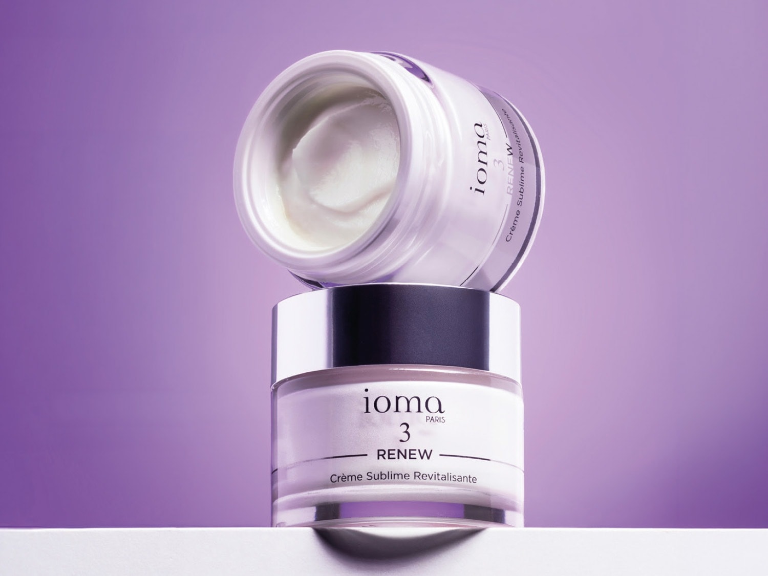 Due vasetti di crema Ioma 3 Renew Crème Sublime Revitalisante sono impilati su uno sfondo viola, con il vasetto superiore aperto che rivela la texture ricca e bianca della crema.