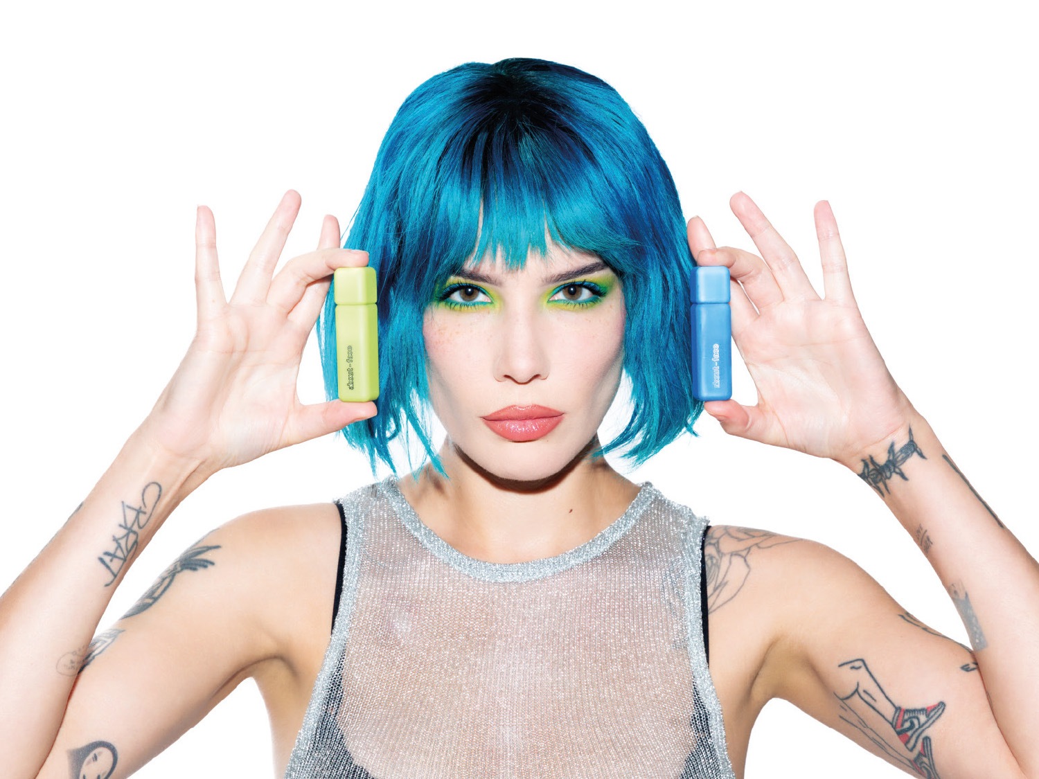 Una donna con capelli blu e trucco verde brillante tiene in mano due piccoli flaconi di profumo, uno verde lime e uno blu, entrambi con la scritta "about-face".