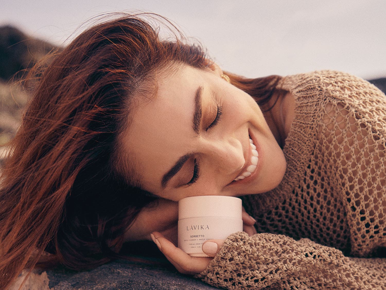 Una donna sorridente con i capelli castani appoggia la guancia su un barattolo bianco di crema Lavika Sorbetto, indossando un maglione a maglie larghe color cammello.
