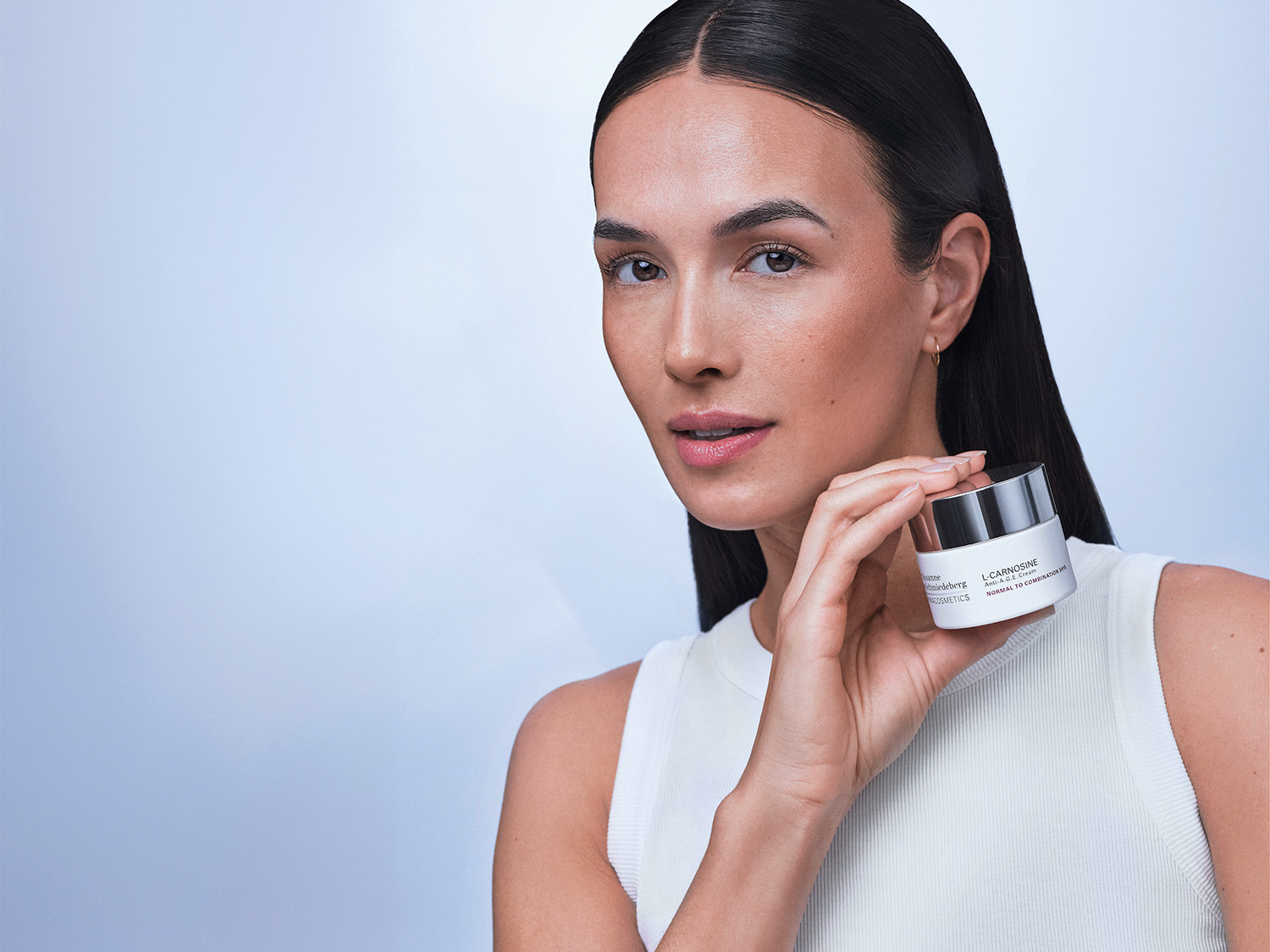 Una donna con lunghi capelli scuri e una canotta bianca tiene in mano un vasetto bianco e argento di L-Carnosine Anti-A.G.E. Crema per pelli da normali a miste.