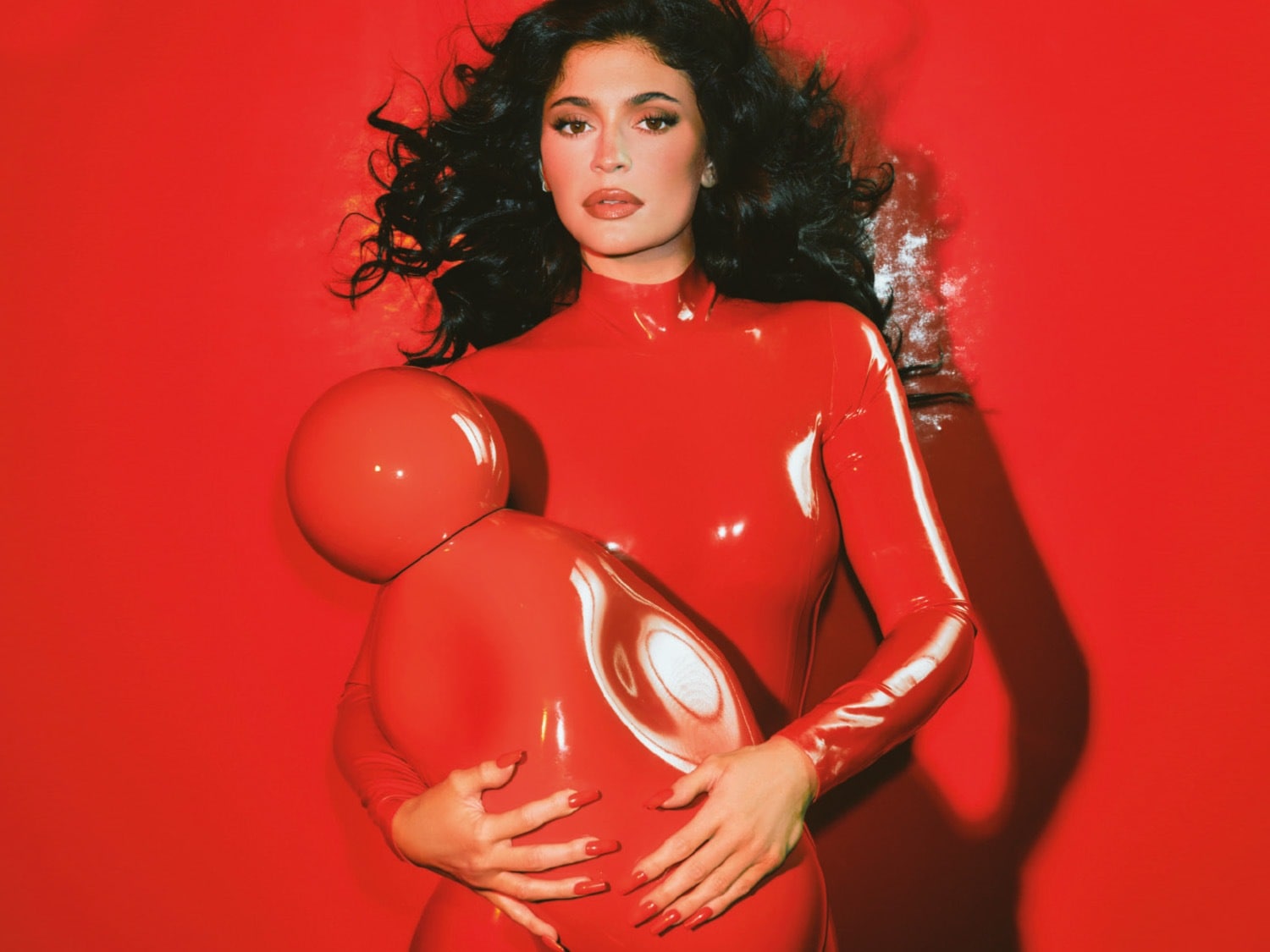 Kylie Jenner indossa una tuta rossa lucida e tiene in braccio una scultura astratta rossa, con i suoi capelli scuri che le incorniciano il viso su uno sfondo rosso vibrante.
