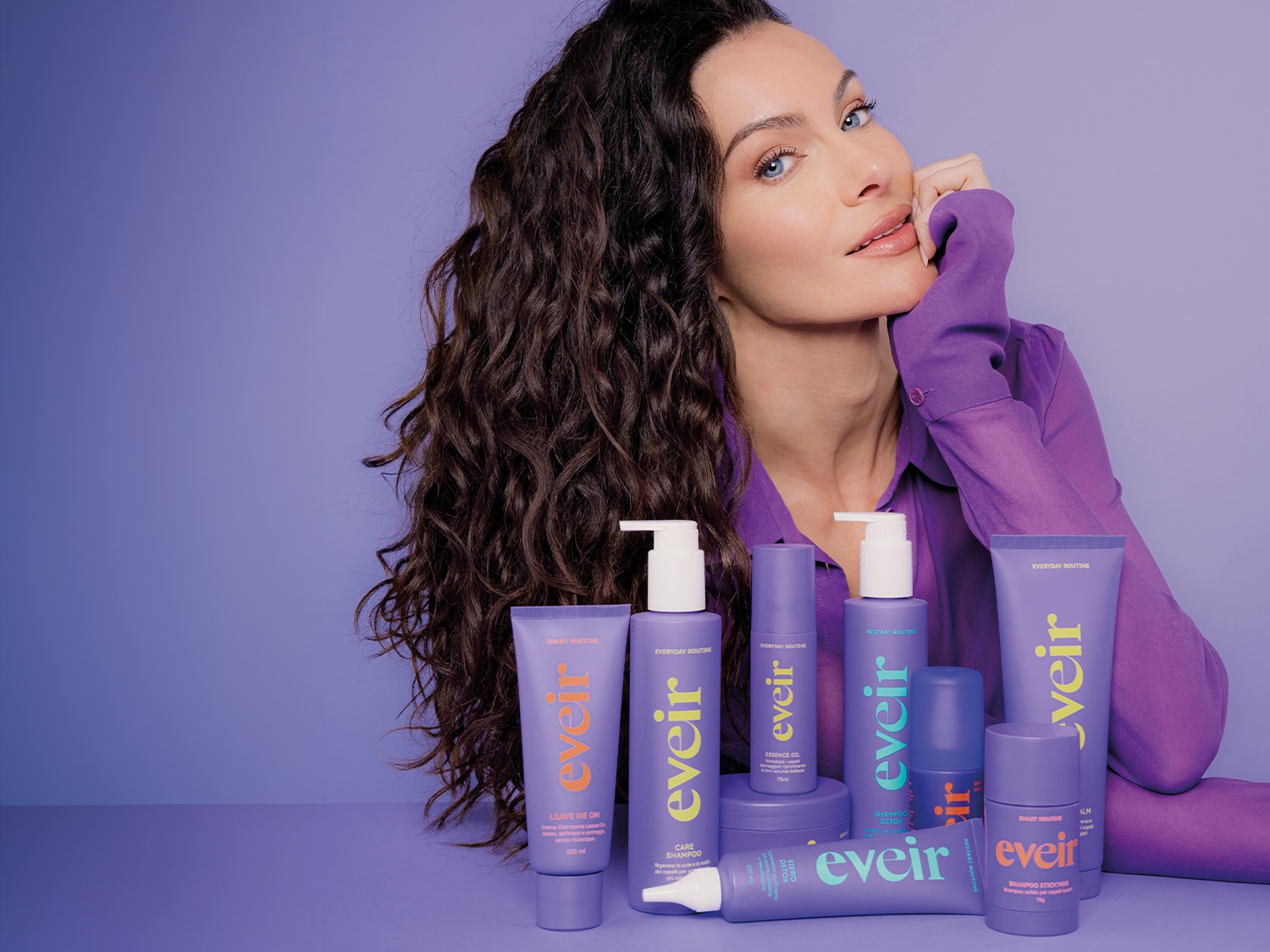 Una donna con lunghi capelli ricci castani e occhi azzurri posa con una mano sul viso, circondata da una collezione di prodotti per capelli viola del marchio "eveir" su uno sfondo viola.