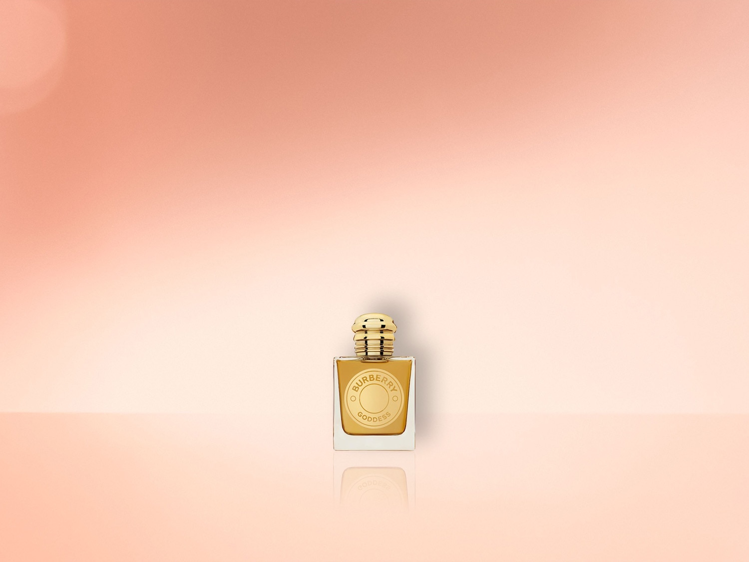 Quadratischer, goldfarbener Burberry Goddess Parfümflakon mit geripptem, kugelförmigem Verschluss auf einem hellpfirsichfarbenen Hintergrund.