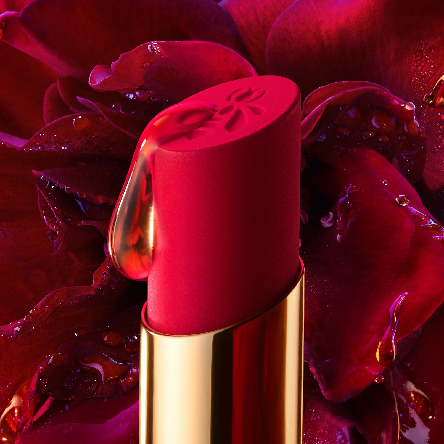 Gros plan d'un rouge à lèvres rouge vif avec un étui doré, orné d'une goutte d'eau brillante, sur un lit de pétales de rose rouge foncé, évoquant une sensation de luxe et d'hydratation.