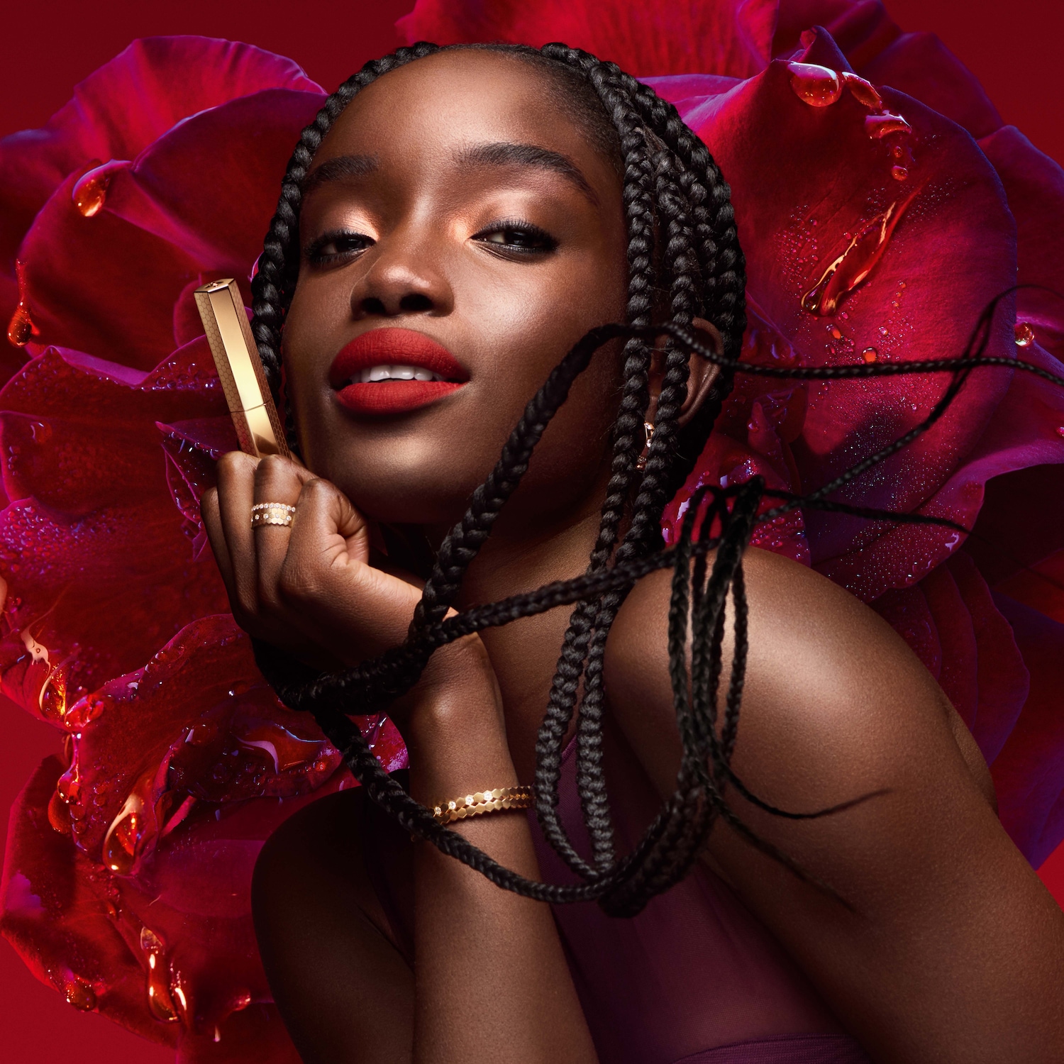 Une femme radieuse, les lèvres rouge vif, tient un rouge à lèvres doré sur un fond de rose rouge luxuriante.