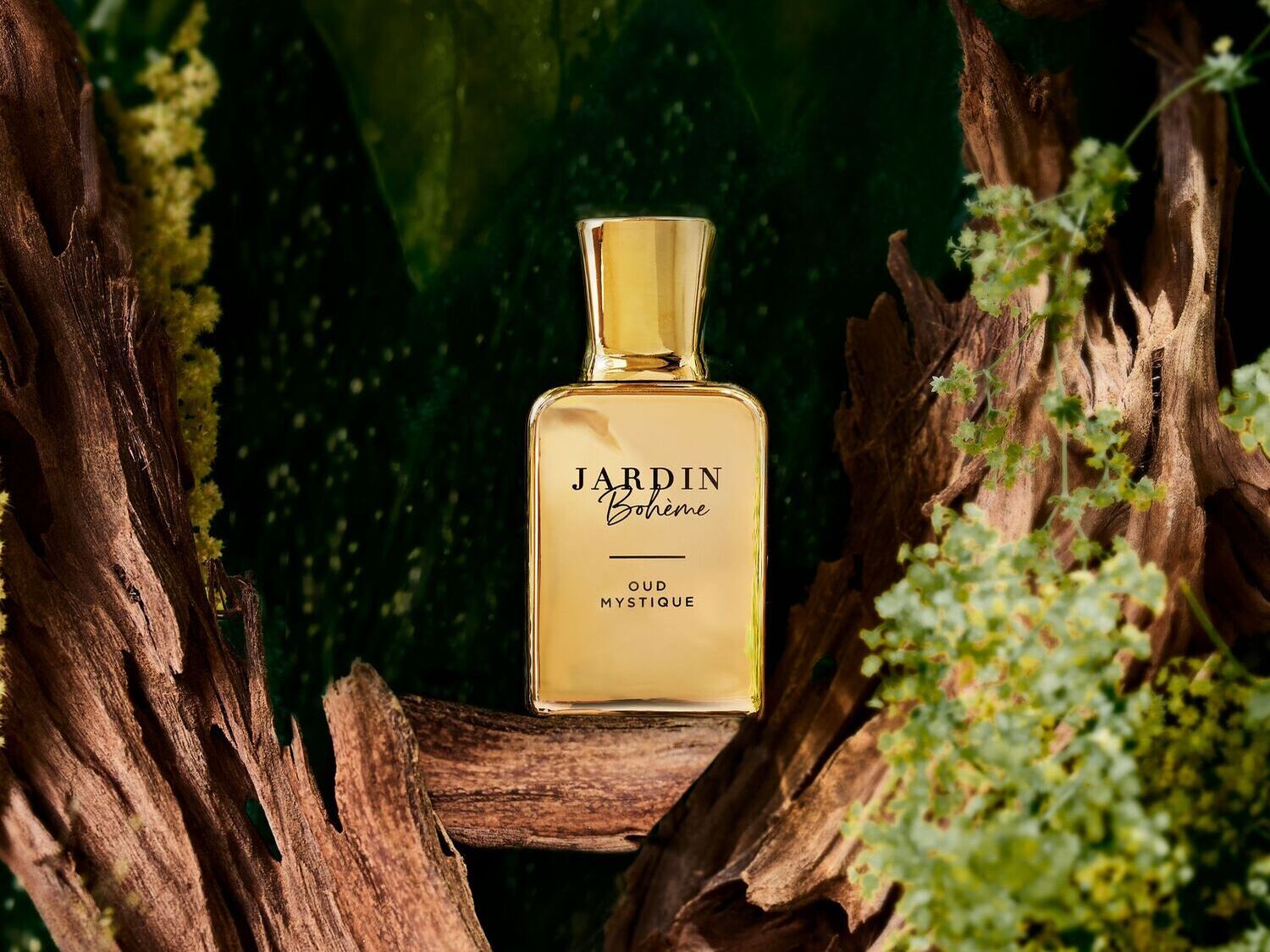 Auksinės spalvos „Jardin Bohème Oud Mystique“ kvepalų buteliukas, apsuptas medienos ir žalios augmenijos, sukuria prabangos ir gamtos harmonijos įspūdį.