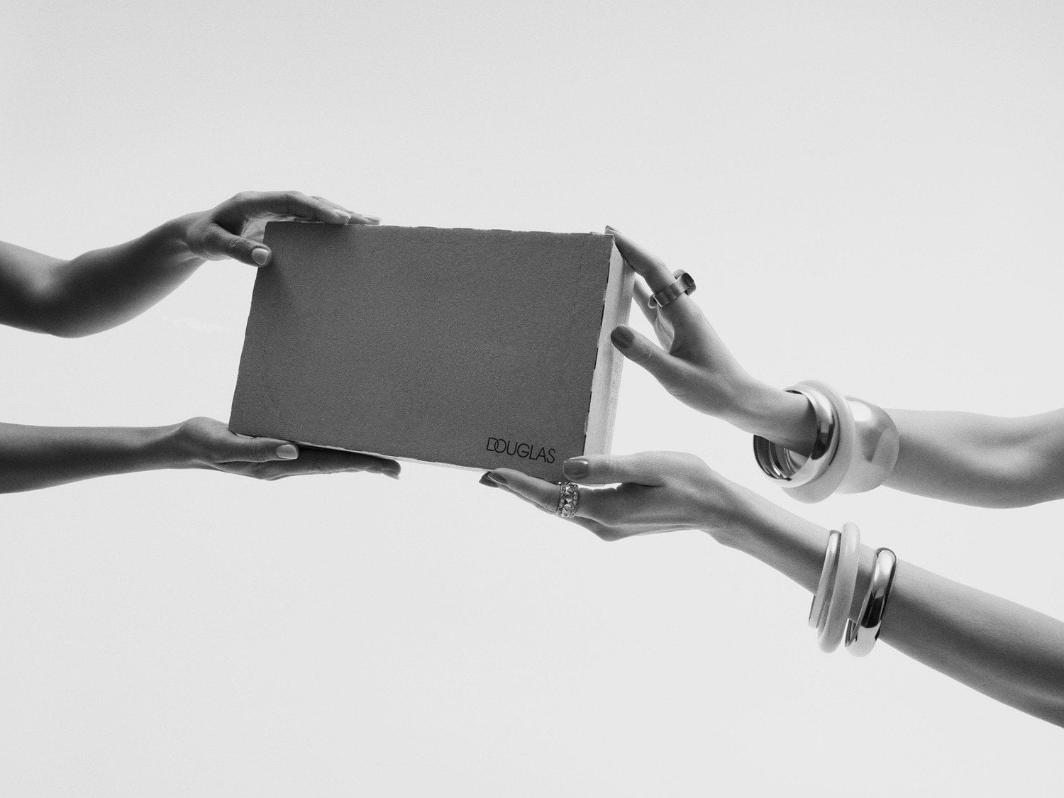 Černobílý snímek zachycuje ruce předávající si balíček od Douglas, zdobené výraznými prsteny a náramky, na světlém pozadí.