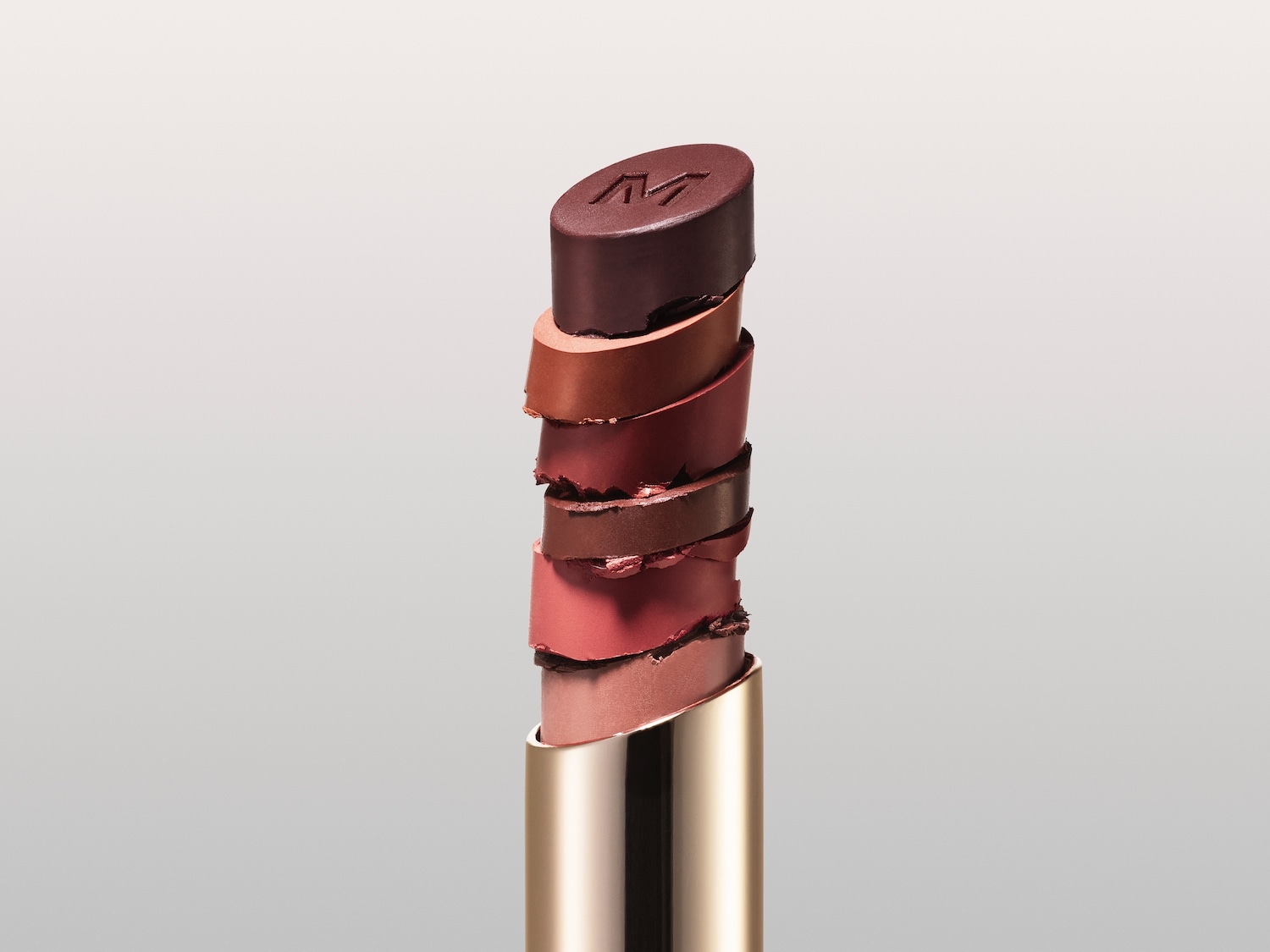 Nahaufnahme eines Lippenstifts mit goldener Hülse, dessen Spitze in verschiedenen Braun- und Rosatönen geschichtet ist und das Markenlogo trägt.