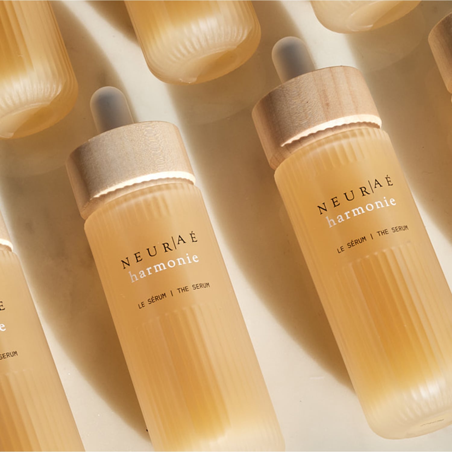 Prezentacja kilku butelek serum NEURAÉ harmonie o pojemności 15 ml, każda z pipetą i drewnianą nakrętką, na kremowym tle.