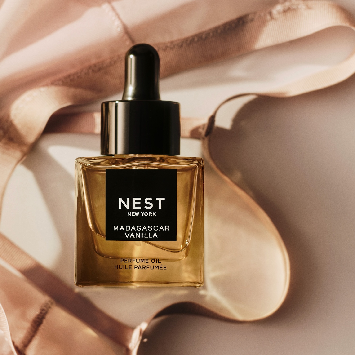„Nest New York Madagascar Vanilla“ kvepalų aliejus yra skaidriame stačiakampiame buteliuke su juodu lašintuvu, o jo auksinis skystis atspindi šviesą, sukuriant šiltą švytėjimą ant šviesiai rožinės liemenėlės.