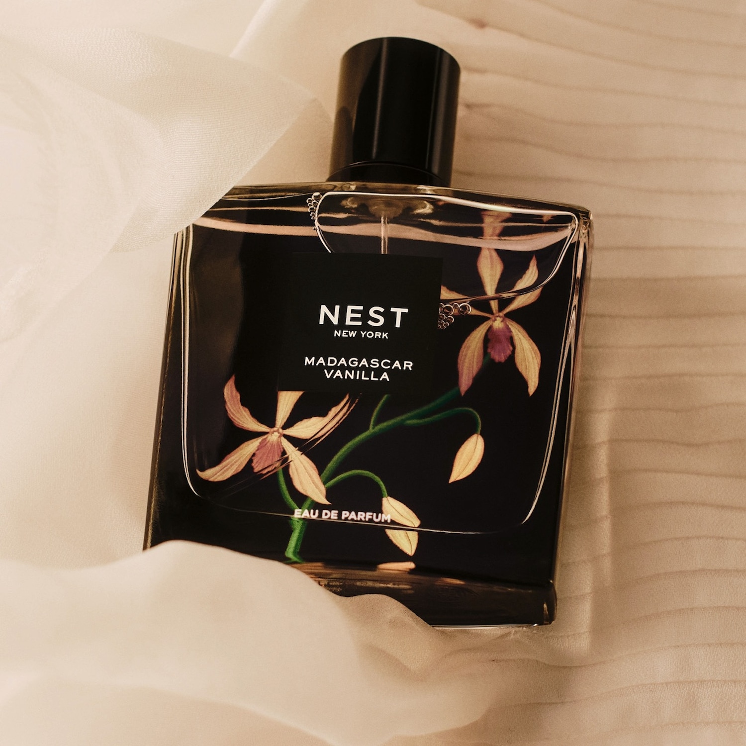 „Nest New York Madagascar Vanilla Eau de Parfum“ kvepalų buteliukas su juoda etikete ir vanilės orchidėjų piešiniais, pastatytas ant šviesaus medinio paviršiaus su balta, permatoma medžiaga.
