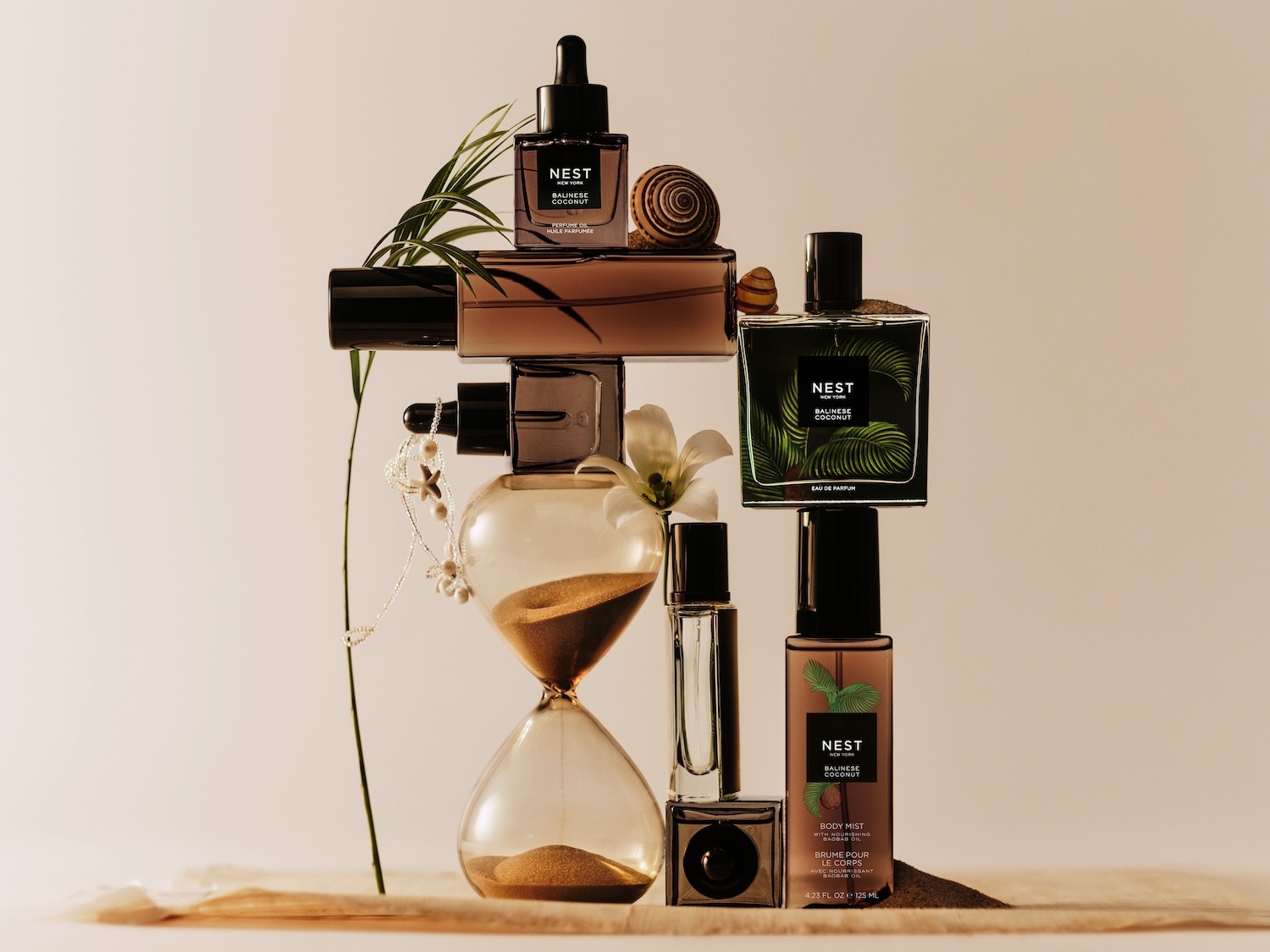 Šiame atvaizde matomas „Nest Balinese Coconut“ kvepalų aliejus, kvepalai ir kūno dulksna, išdėlioti ant smėlio laikrodžio, apsupti kriauklių ir augalų, kurie sukuria atpalaiduojančią ir egzotišką atmosferą.