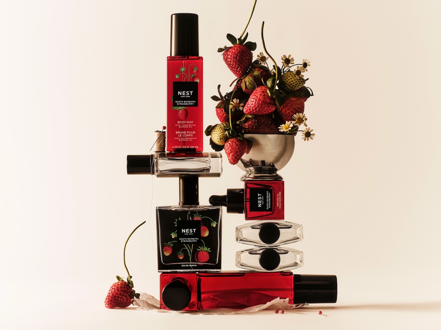 NEST Santa Barbara Strawberry kūno dulksna raudoname buteliuke su juodu dangteliu, papuošta braškių piešiniais, stovi ant kitų NEST produktų, o šalia - dubenėlis su šviežiomis braškėmis ir ramunėlėmis.