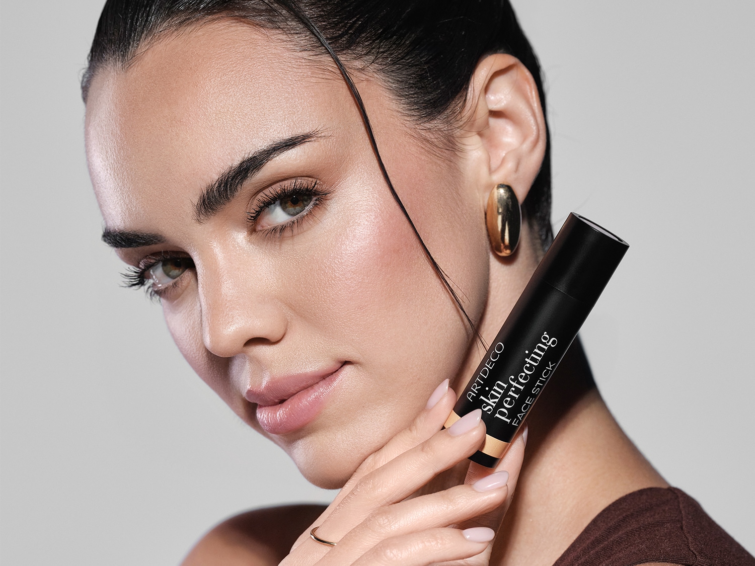 Eine Frau mit braunen Haaren und goldenen Ohrringen hält einen schwarzen Skin Perfecting Face Stick von ARTDECO in der Hand, der einen hellen Farbton hat.