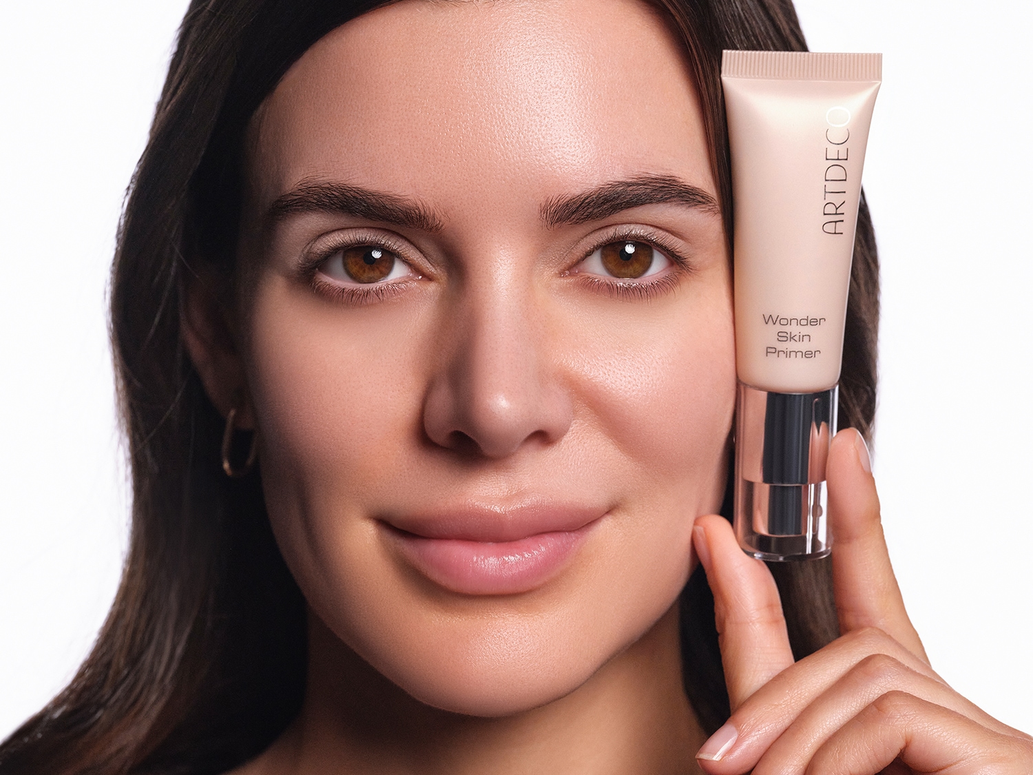 Eine Frau mit braunen Haaren hält eine Tube ARTDECO Wonder Skin Primer neben ihr Gesicht.