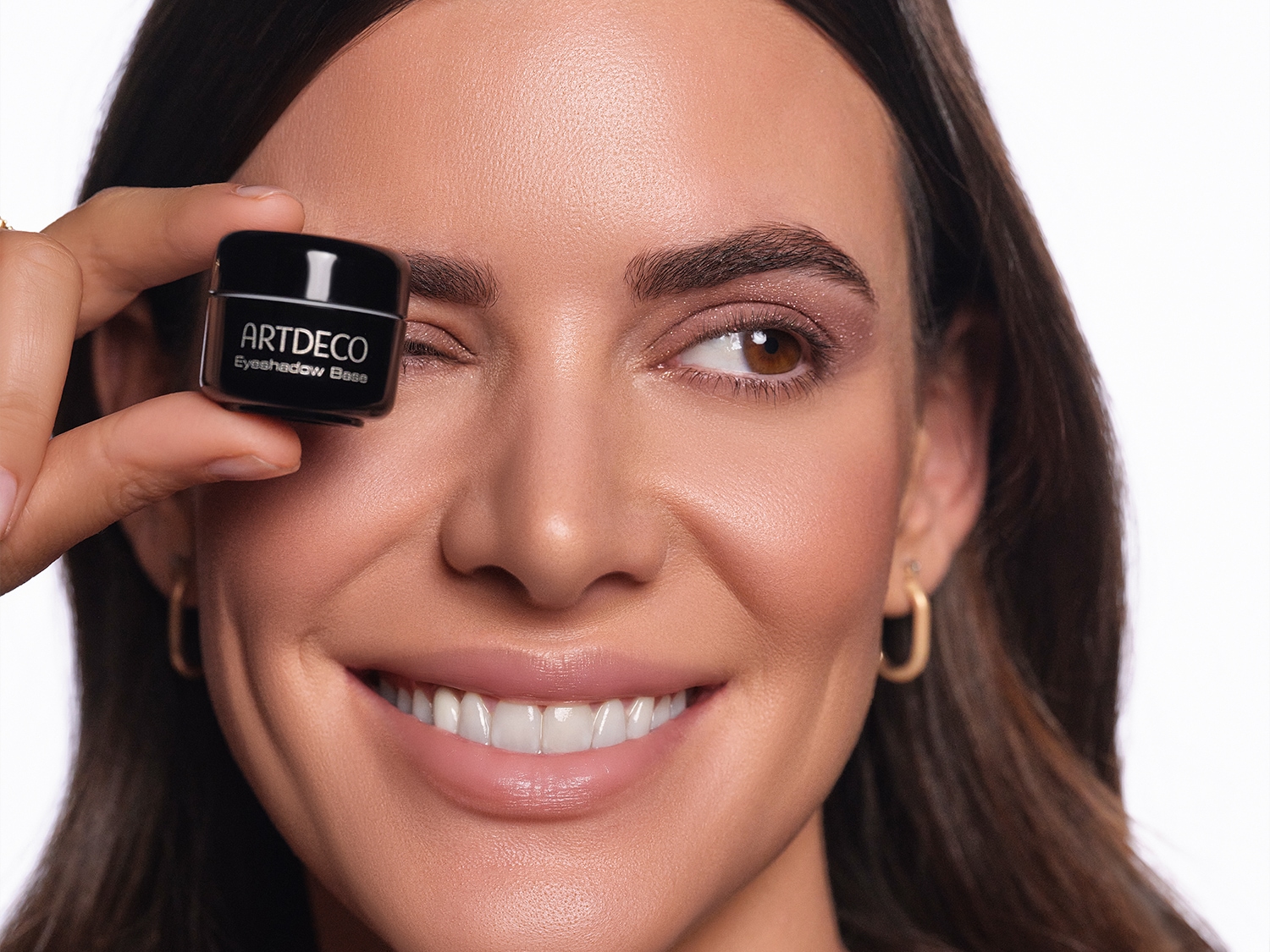 Eine lächelnde Frau hält einen kleinen, schwarzen Tiegel mit der Aufschrift ARTDECO Eyeshadow Base vor ihr rechtes Auge.