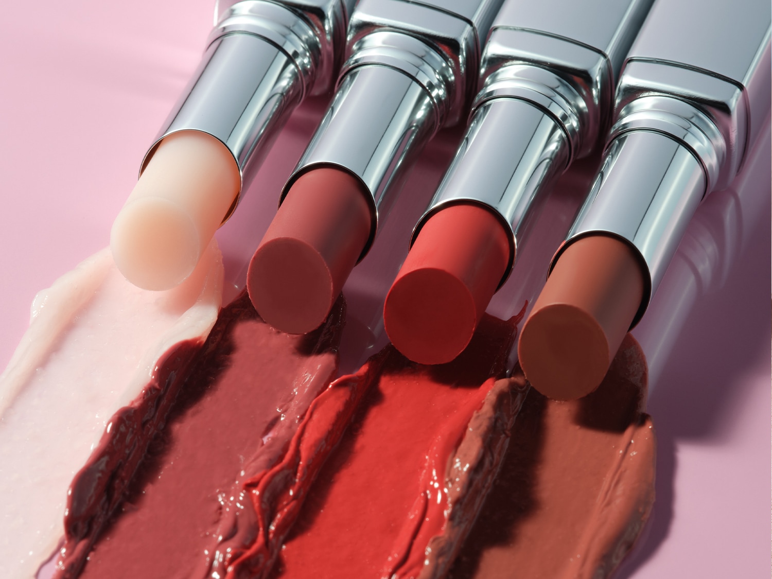 Nahaufnahme von vier Lippenstiften in verschiedenen Farbtönen, von blassrosa bis braun, mit jeweiligen Farb-Swatches auf einem rosafarbenen Untergrund.