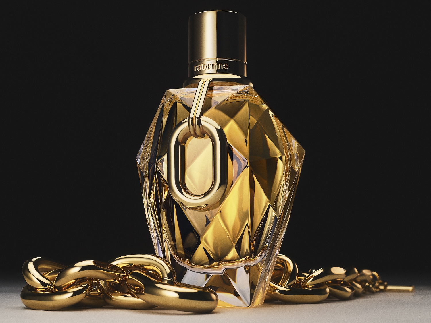 Złoty flakon perfum Rabanne FAME o fasetowanym kształcie, przypominającym klejnot, z ozdobnym łańcuchem i złotą zawieszką, leży na białej powierzchni na ciemnym tle.