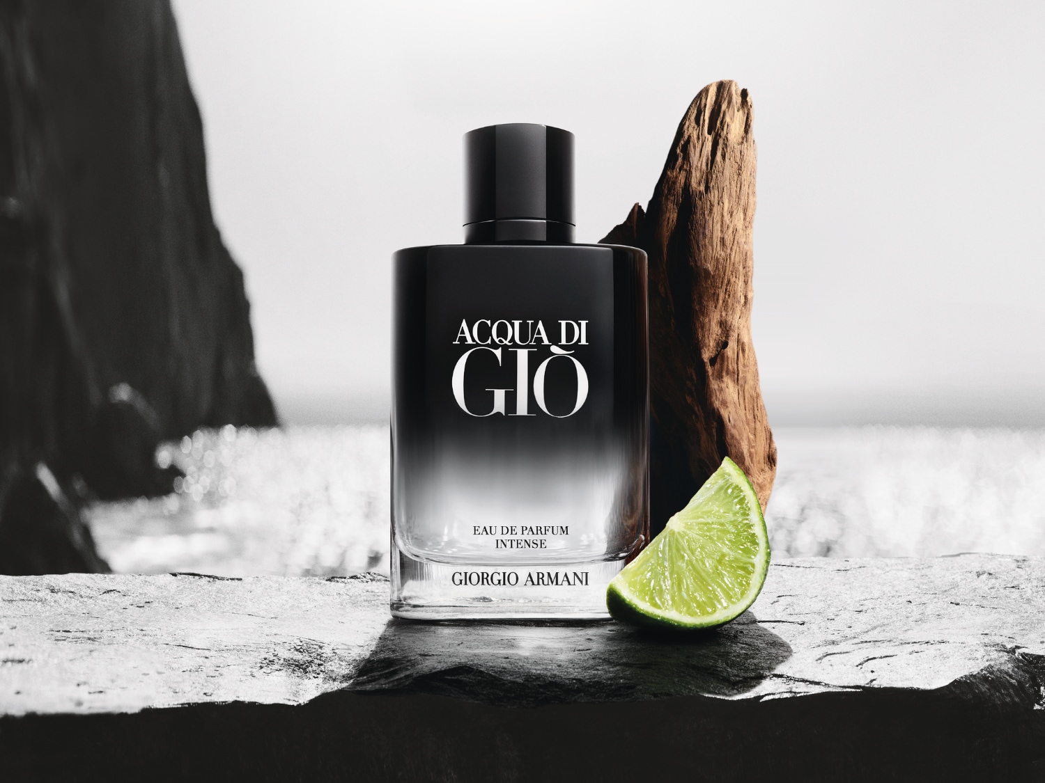 Flakón Acqua di Giò Eau de Parfum Intense od Giorgia Armaniho s čiernym vrchnákom, drevom a limetkou na pozadí mora.