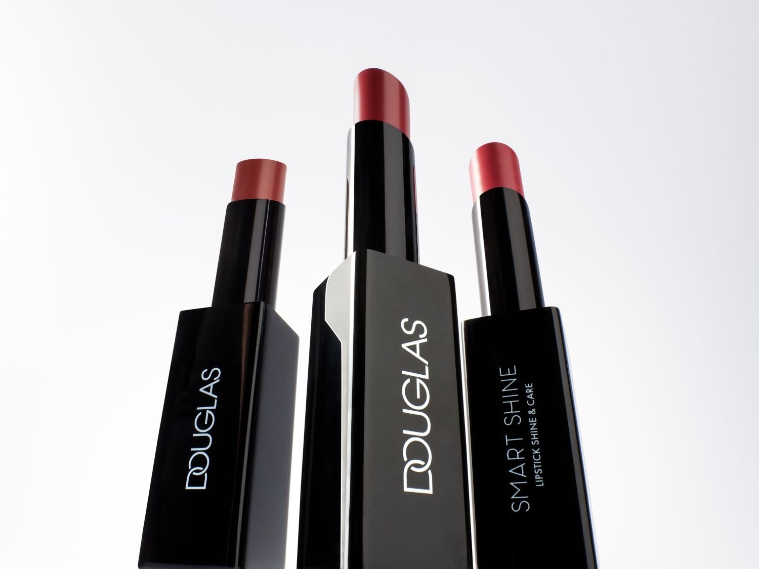 Drei Douglas Smart Shine Lippenstifte in ihren schwarzen Hüllen mit rotem Lippenstift, der oben herausschaut, versprechen Glanz und Pflege.