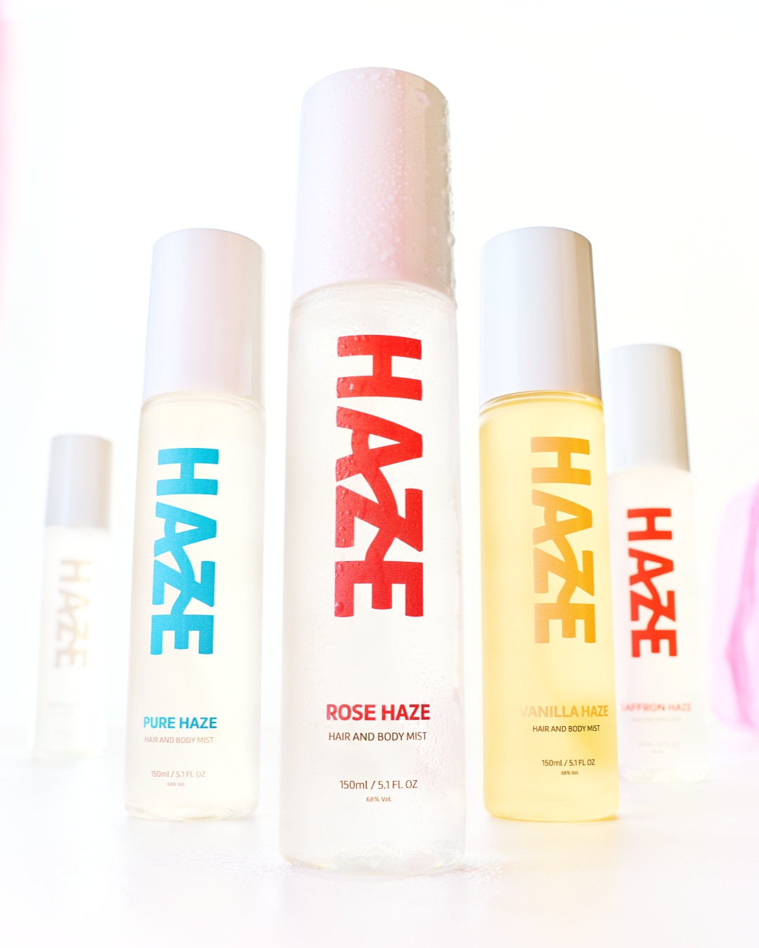 Esplora la collezione Haze di spray per capelli e corpo: Pure Haze in flacone trasparente con scritta blu, Rose Haze con scritta rossa, Vanilla Haze con liquido giallo e Saffron Haze.