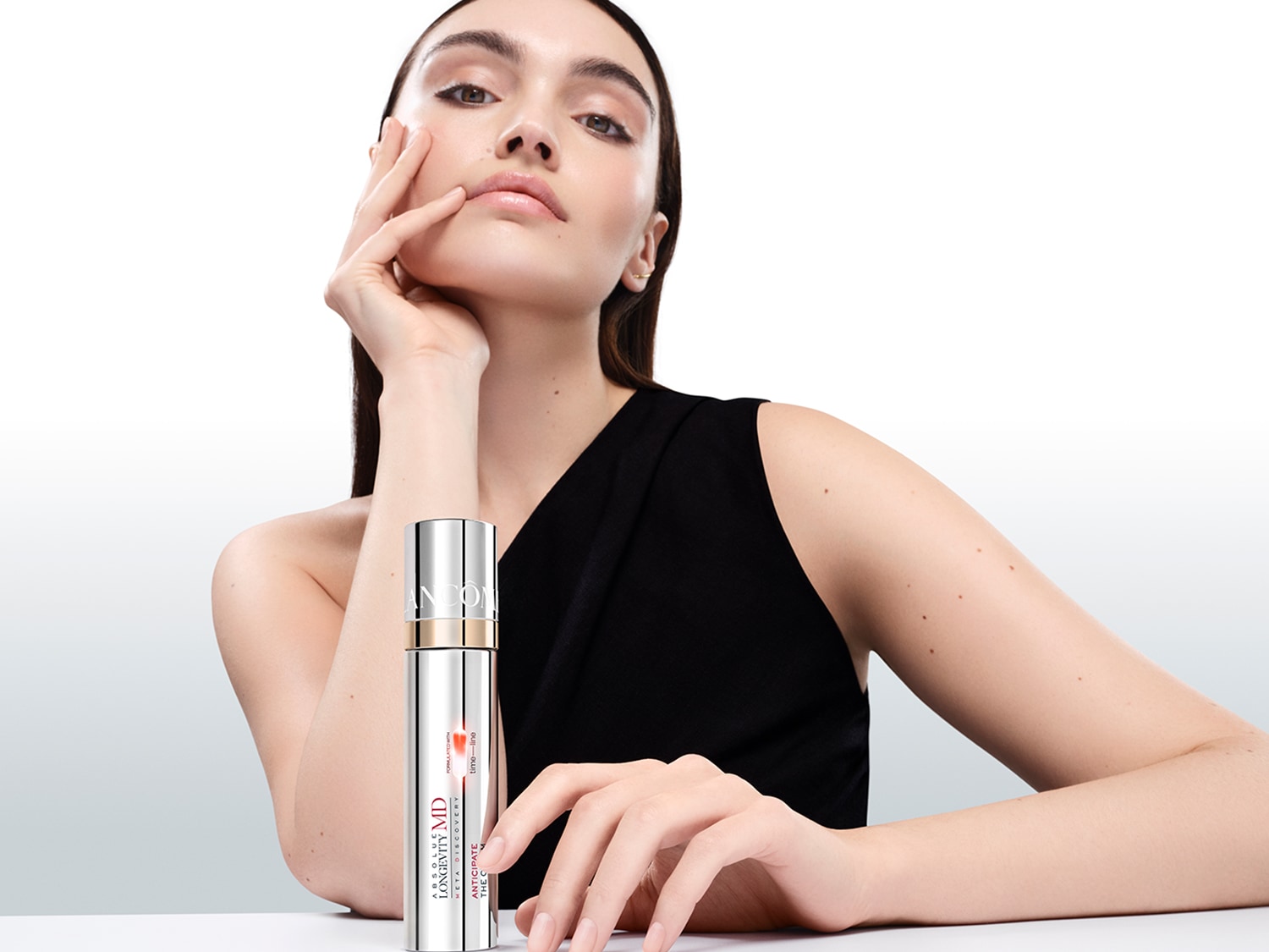 Elegantiška moteris su juoda suknele pozuoja su „Lancome Longevity MD“ serumu sidabriniame buteliuke, pabrėždama odos priežiūros priemonės prabangą.
