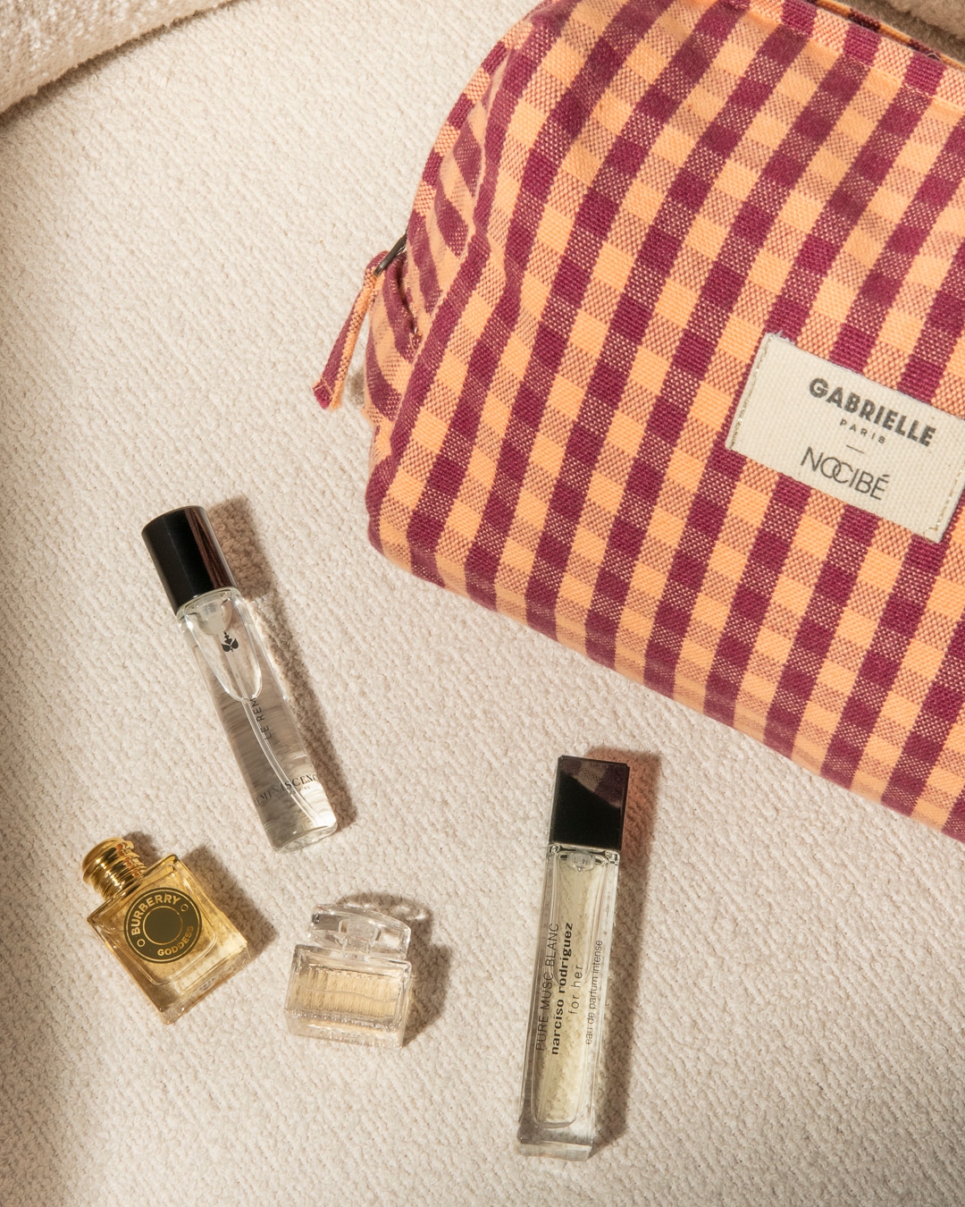 Découvrez un assortiment de parfums miniatures, présentés à côté d'une trousse à carreaux vichy orange et bordeaux, idéale pour vos voyages.