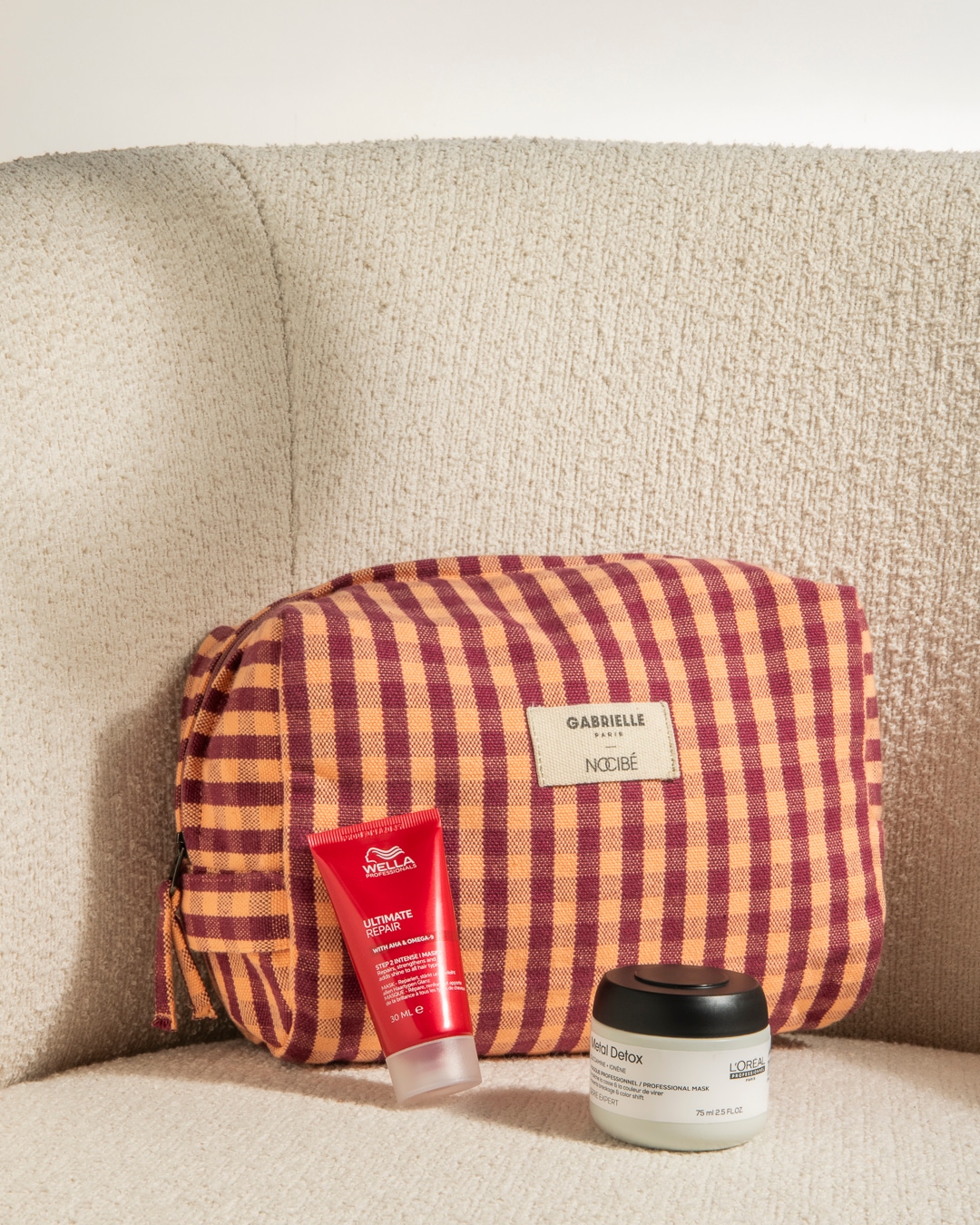 Trousse à carreaux orange et bordeaux Gabrielle Paris Nocibé, avec un tube rouge Wella Ultimate Repair et un pot blanc L'Oréal Metal Detox, posés sur un fauteuil beige.