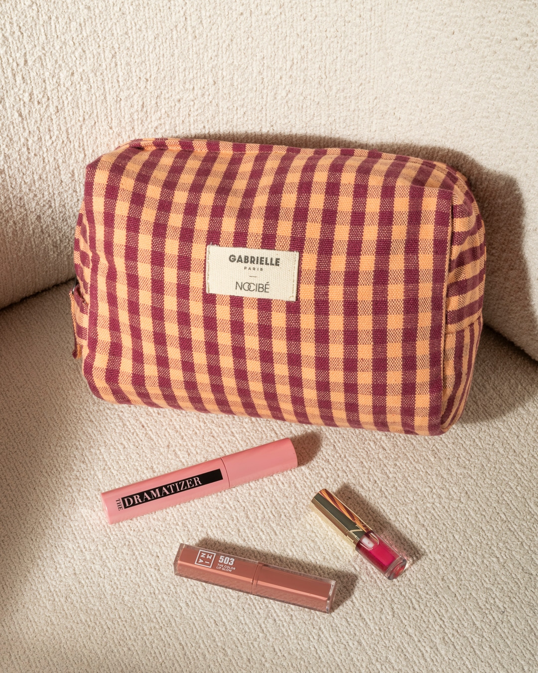 Voici une trousse à maquillage à carreaux orange et bordeaux, accompagnée d'un mascara rose Dramatizer et de deux brillants à lèvres.