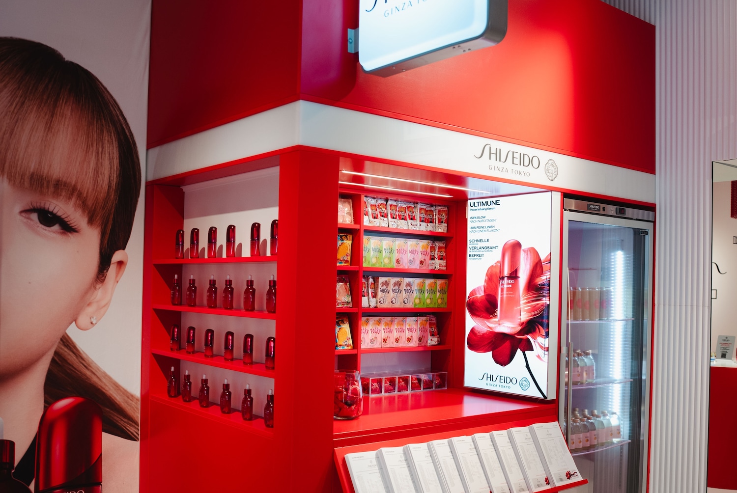 Ein roter Shiseido-Stand mit Regalen voller Hautpflegeprodukte, darunter Ultimune Power Infusing Serum, Pocky und eine beleuchtete Anzeige mit dem Slogan „+54 % Glow nach nur 3 Tagen“.