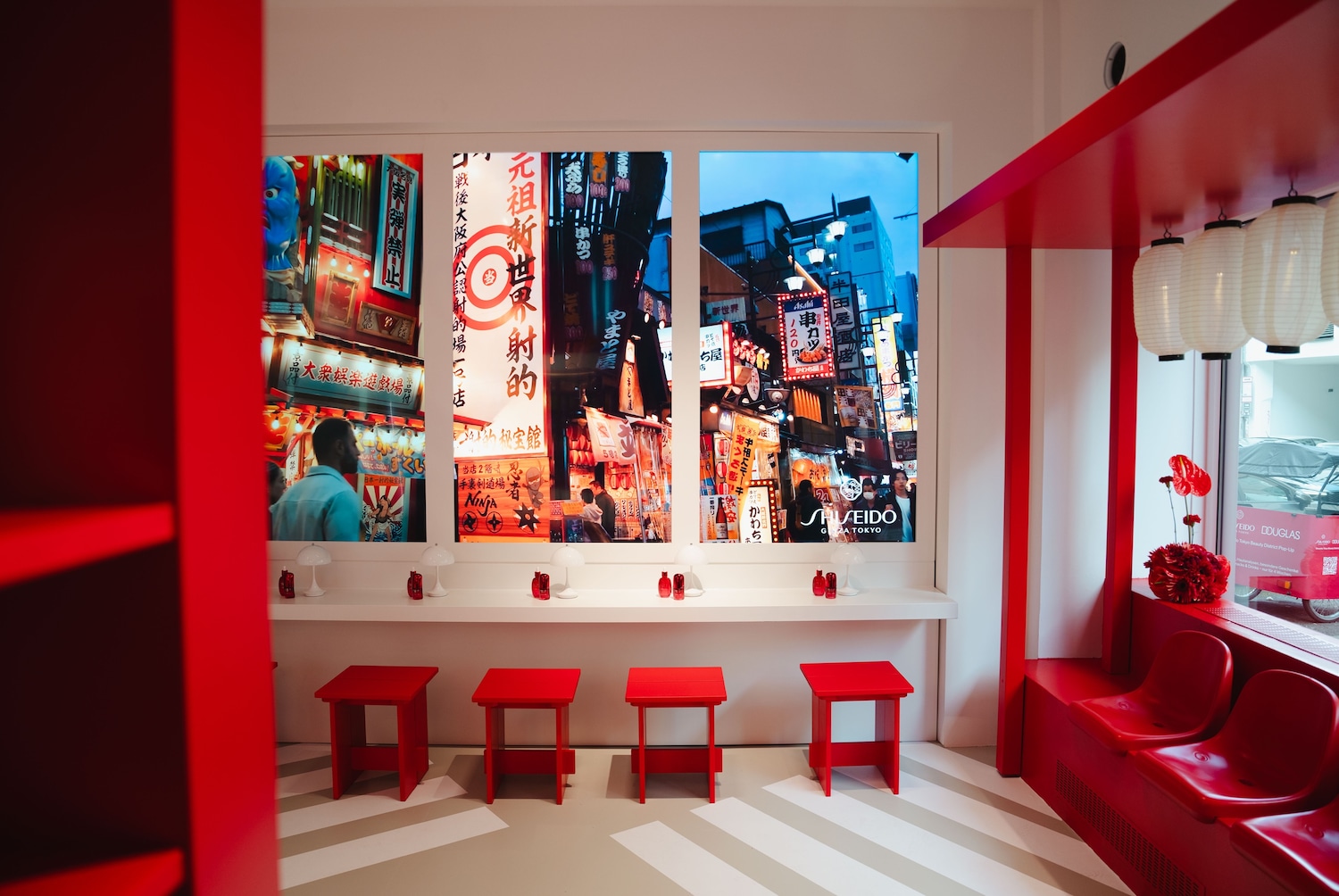 Einladender Wartebereich in Rot und Weiß mit Blick auf eine belebte japanische Straßenszene, die durch Fenster simuliert wird, die mit Shiseido-Produkten dekoriert sind.