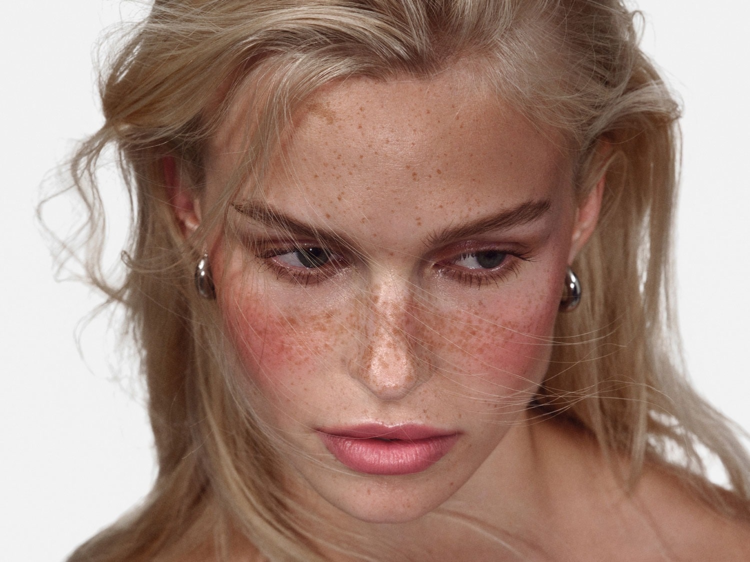 Close-up van een vrouw met blond haar, sproeten en een lichte blush op haar wangen, kijkend naar beneden.