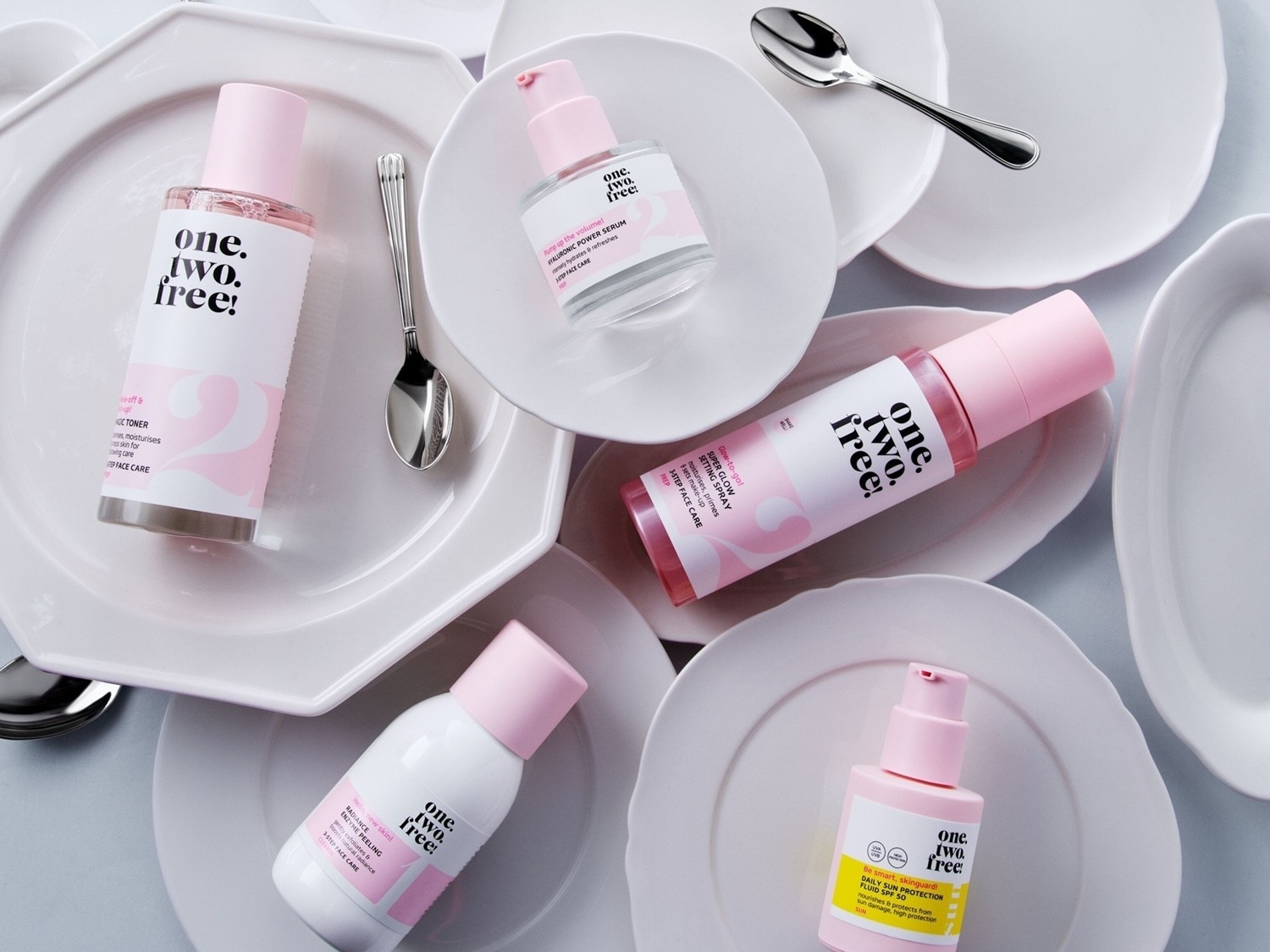Descubre la colección One.Two.Free! con tónico, sérum, peeling enzimático, spray fijador y fluido de protección solar SPF 50, presentados en envases blancos y rosas sobre platos blancos.